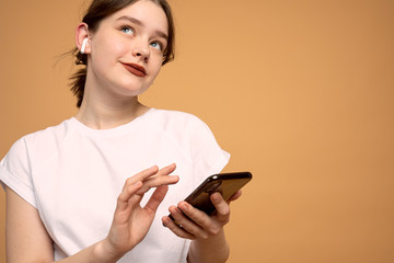 Young business lady in white t-shirt holding mobile phone in hands, wearing wireless headphones, looking up, isolaetd over orange background