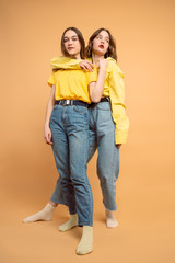 Full length of two cute confident young women in yellow shirts hugging over orange background