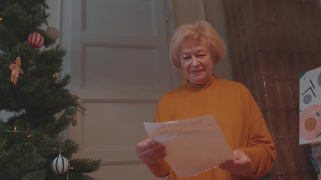 Infant baby child curiously playing near Christmas tree. His granny beautiful mature woman looking at painting staying at home happy for holidays.