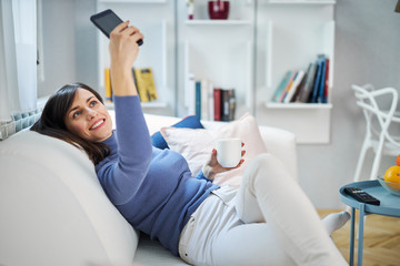 Young charming cheerful smiling brunette sitting on couch in her loving room, holding mug with fresh coffee and taking selfie.