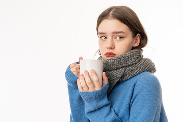 Flu treatment. Portrait of sick brunette teenage girl wearing casual clothes and scarf, holding cup of tea, studio shot isolated on white background