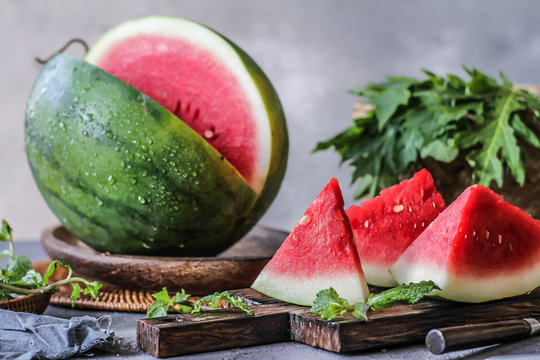 Photo of fresh watermelon with green leaves on wooden board over the table. Whole and sliced. Cutting board. Cut in halves. Dark background. Front view. Raw organic fruit. Image