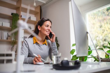 Attractive businesswoman sitting at the desk indoors in office, using smartphone.