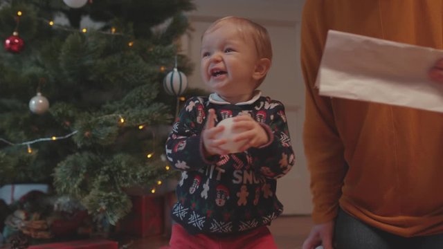 Cheerful granny upbringing her little grandchild playing with baby boy near decorated Christmas tree at home on holidays. Family reunion.