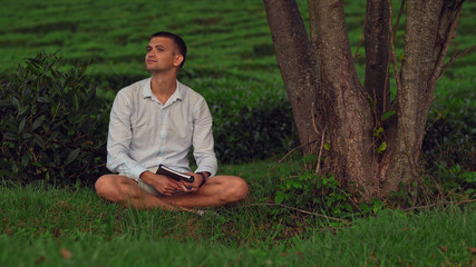 A young man sits on the grass under a tree with a notebook and enjoys nature. Tea plantations in...