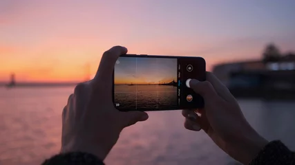 Selbstklebende Fototapeten Hellrosa Nahaufnahme der Hände einer Frau mit einem Telefon, das Fotos eines hellen Sonnenuntergangs und des Meeres macht.  © StudioPN