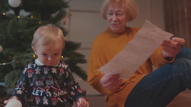 Beautiful happy grandmother playing with infant grandchild showing a painting talking to cute baby staying together near Christmas tree at home.
