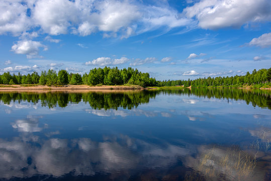 Finland, Lapland, Ivalo: Panorama view of pure rural Finnish nature with calm lake water, cloud reflections, green trees, riverbank and blue sky in background - concept remote rural nobody environment