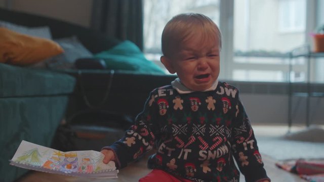 Poor naughty young toddler baby boy crying with tears holding a paper drawing look at camera in the house room.