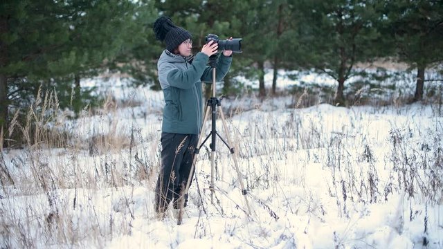 The girl enthusiastically takes pictures in the winter forest on a photo camera, which is standing on a tripod, general view