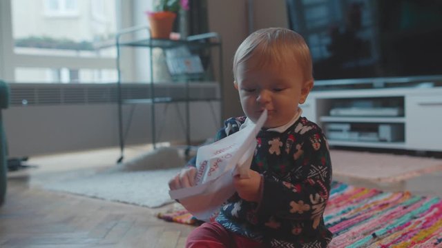 Lovely curious baby boy in holiday sweater crawling on floor in the room playing and squeezing paper drawing look at camera. Family and childhood concept.