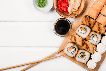 Japanese sushi rolls set with salmon acne and cream cheese on bamboo board with chopsticks and fresh food ingredients with soy sauce and ginger on white wood copyspace background.