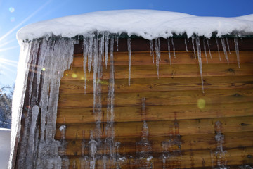 The bright spring sun shines on the snow and icicles sparkle on the wooden wall. Icicles descend from the roof of a wooden house and reflect the rays of the sun.