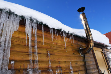 The sun shines brightly, early spring icicles melt. A tin pipe sticks out of a wooden house, the rays of the sun are reflected on the pipe. Icicles hanging from the roof of a wooden house. © atevi
