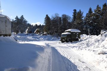 Fototapeta na wymiar Snow-covered terrain, snow and forest, and in the distance a barrier is visible, behind it - a large tractor in the snow and several camp guests' cars. Military car in the snow.