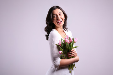 Studio portrait of gorgeous young brunette woman with long wavy hair wearing tight white wedding dress , holding bouquet of pink tulip flowers. Grey isolated background, copy space, close up.