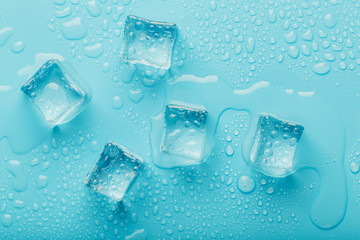 Ice cubes with water drops scattered on a blue background, top view.