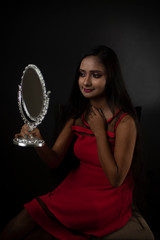 Fototapeta na wymiar Fashion portrait of an young and attractive Indian Bengali brunette girl in red western dress holding a mirror in her hand in front of a black studio background. Indian lifestyle
