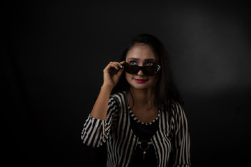 Obraz premium Fashion portrait of an young Indian Bengali brunette girl with striped western semi formal/casual dress with sunglasses in front of a black studio background. Indian fashion portrait and lifestyle.
