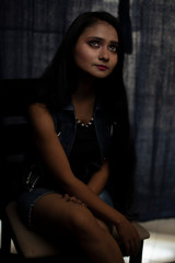Fashion portrait of an young and attractive Indian Bengali brunette girl with tee shirt and blue western jacket sitting in front of a black studio background. Indian fashion portrait and lifestyle.