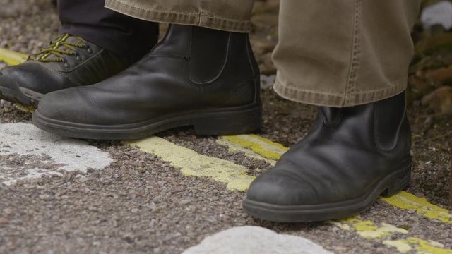 A point-of-view, closeup shot of a pair of feet wearing leather boots and pants standing on the painted yellow lane indicator of a driveway.