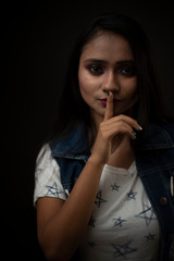 Obraz premium Fashion portrait of an young Indian Bengali brunette girl with tee shirt and blue western jacket putting finger on lips in front of a black studio background. Indian fashion portrait and lifestyle.