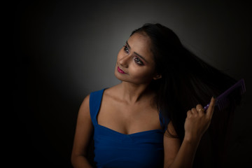 Fototapeta premium Fashion portrait of young Indian Bengali brunette girl with blue western dress combing her hair looking at mirror in front of black studio background. Indian fashion portrait and lifestyle.