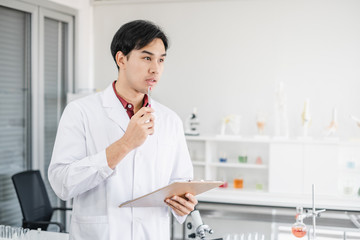 A male asian scientist writing on a clipboard in white laboratory with test tube microscope and solutions.