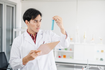 A male asian scientist holding a test tube with blue solution in one hand and clipboard in another...