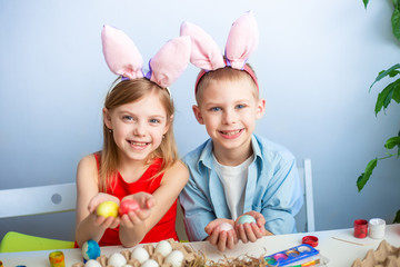 cute smiling children, fair-haired brother and sister of 7-9 years old, wear bunny ears on their heads and paint Easter eggs on a blue background