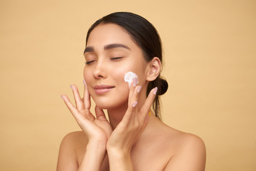 Young girl with closed eyes with clean skin puts cream on her face with hands on a yellow background