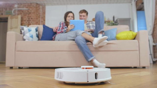 Young husband and wife resting with tablet while automatic vacuum cleaner cleaning floor