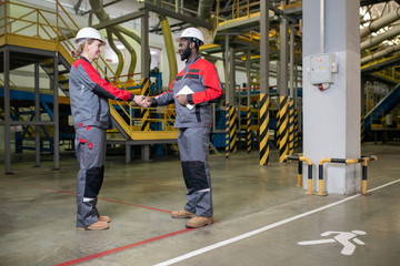 Horizontal shot of two factory engineers wearing uniform greeting each other with handshake