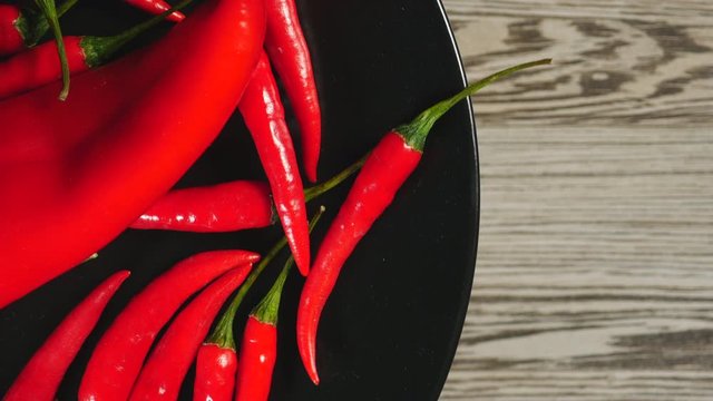 Top View Of Fresh Raw Red Chili Peppers And Ramiro Pepper Rotating On A Black Plate On A Gray Wooden Background On The Left Side.