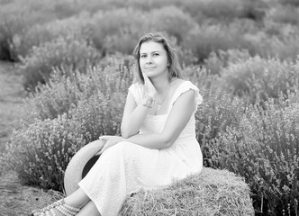 woman in a white sundress sits on a haystack in a lavender field and rests. Holding a straw hat