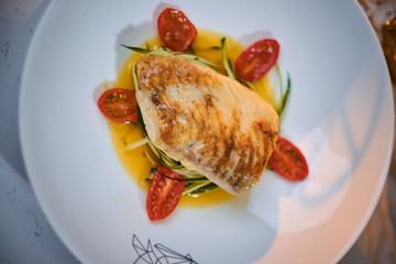 Delicious fillet of grilled and oven baked codfish or coalfish served with thin sliced vegetables and oil sauce, on a white porcelain dish next to golden candles in a warm light of a restaurant, close