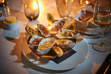 Rich and luxury dinner golden metallic bowl of prepared oysters and crispy bread loafs with sauce and vegetable spread served on a white stone table next to candle, a glass of white wine, jellyfish in