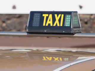 A taxi sign has a green light indicating it is available for hire.It is attached to the vehicles roof bar.It is reflected in the cars roof.Image