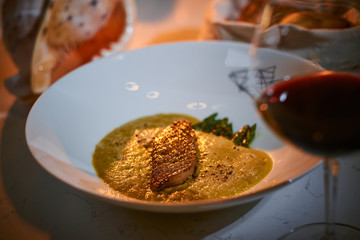 Excellent fillet of grilled and oven baked gourmet fish served with garnish including herbs and oil sauce, on a white porcelain dish next to big seashell, a glass of red wine and a full wicker basket