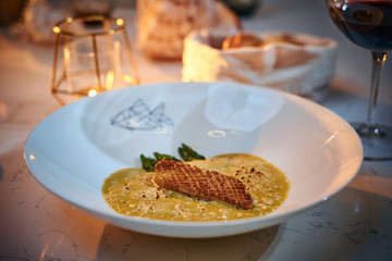 Excellent fillet of grilled and oven baked gourmet fish served with garnish including herbs and oil sauce, on a white porcelain dish next to golden candle, a glass of red wine and a full wicker basket
