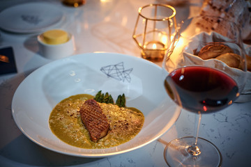 Haute cuisine fillet of grilled and oven baked gourmet fish served with garnish including herbs and oil sauce, on a white porcelain dish next to golden candle, a glass of red wine and a full wicker