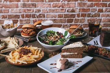 View of the table with hamburgers, french fries and salad, drinks and cake and desserts on the wooden table.