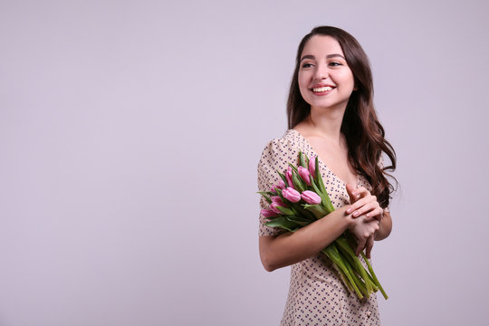 Studio portrait of gorgeous young brunette woman with long wavy hair wearing slim cotton dress, holding bouquet of pink tulip flowers. Grey isolated background, copy space, close up.