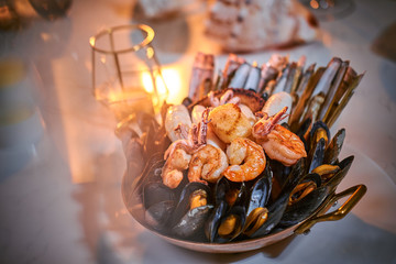 A full dinner rich ceramic bowl of prepared grilled shrimps and mussels including tiny octopus served on a white stone table next to golden candle in a soft light of a restaurant, close up