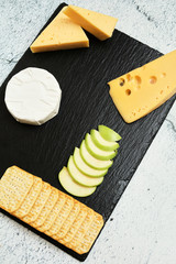 Pieces of cheese on a dark background. Cheeseboard. Sliced ​​apple and nuts on the board. Hard cheese, camembert, roquefort, emmental.