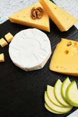Pieces of cheese on a dark background. Cheeseboard. Sliced ​​apple and nuts on the board. Hard cheese, camembert, roquefort, emmental.