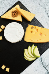 Pieces of cheese on a dark background. Cheeseboard. Sliced ​​apple and nuts on the board. Hard cheese, camembert, roquefort, emmental.