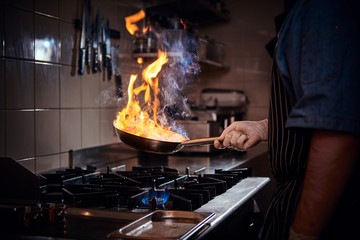 Professional cook wearing gloves and apron cooking stir-fry flambe, creating flames, holding a pan with one hand in a dark restaurant kitchen