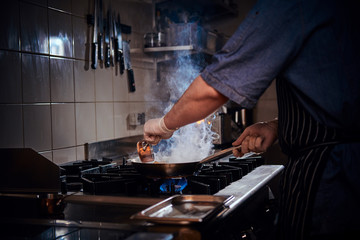 Professional chef wearing gloves and apron mixing and frying stir-fry flambe on a pan with lots of steam in a dark restaurant kitchen