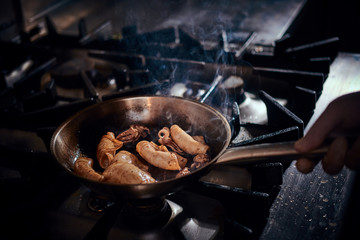 Person wearing gloves cooking stir-fry seafood with squids and fish pieces on a pan with steam in a dark restaurant kitchen